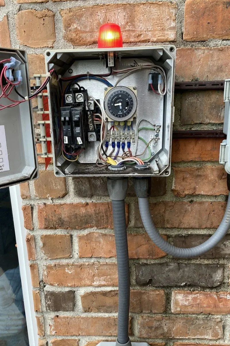 Testing the control panel behind a septic alarm: a red beacon light on top and an open panel with a clamp meter reading the wiring