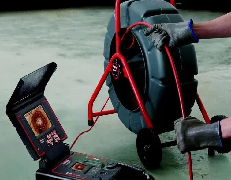 Push camera reel with red cable being fed by a technician in work gloves, with the monitor showing the inside of a pipe
