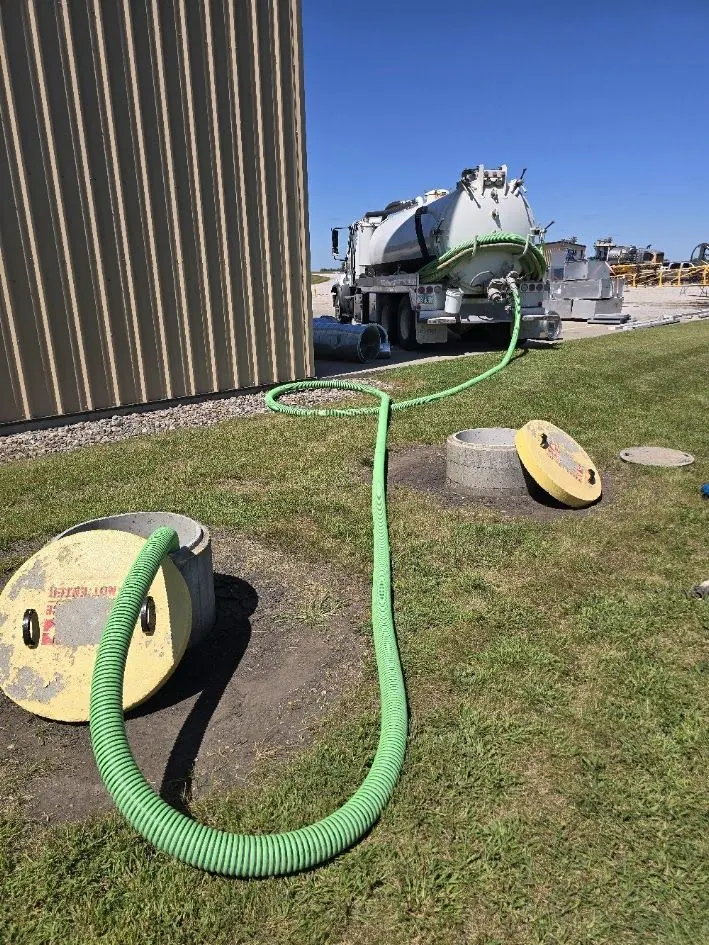 Vacuum truck pumping out a septic system through a green hose, with two concrete riser openings and their yellow lids lying on the grass