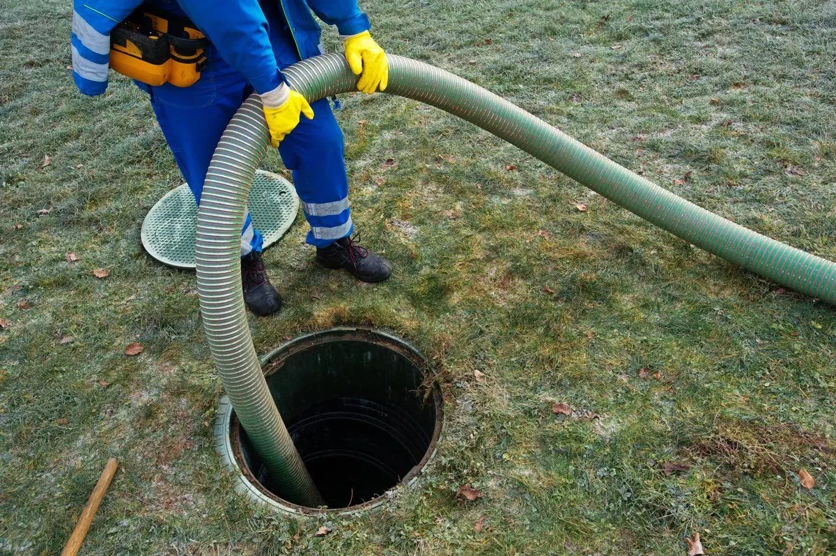 Technician in blue coveralls and yellow gloves guiding a vacuum hose down an open septic tank access on a lawn, with the lid set aside