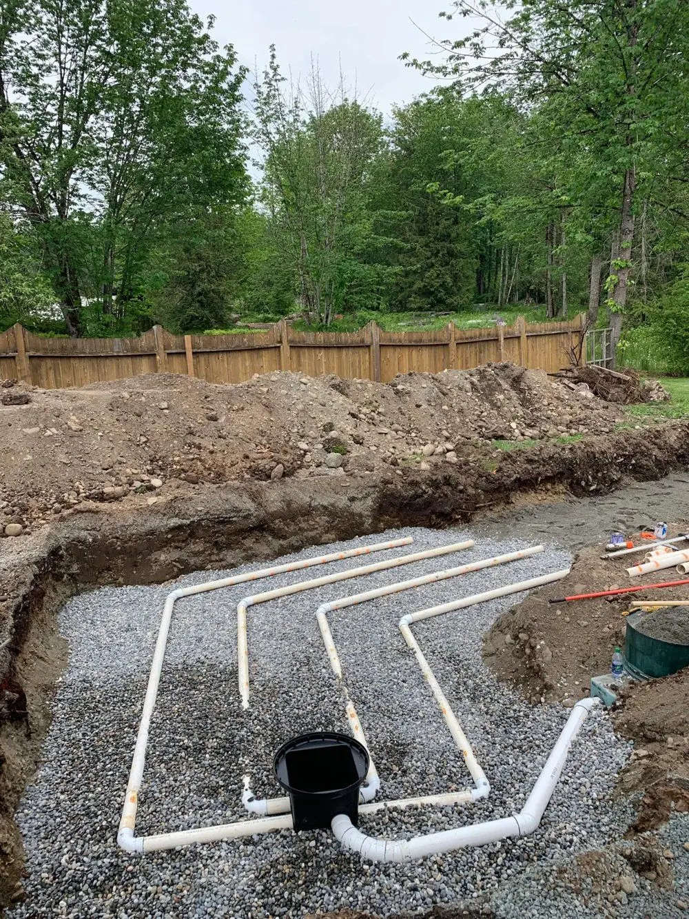 New septic drain field laid out in an excavation: white perforated pipes on a bed of washed gravel, fed from a black distribution box